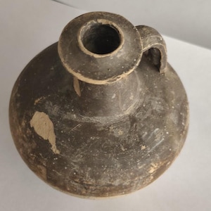 Puede incluir: Una jarra de cerámica marrón oscuro de estilo antiguo, con cuerpo redondeado, asa pequeña y abertura circular ancha. La jarra tiene un aspecto desgastado con desgaste visible, incluyendo áreas astilladas, lo que sugiere edad y uso.