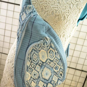 K&ouml;nnte beinhalten: Eine hellblaue Sling-Bag mit geometrischem Muster und Rei&szlig;verschluss. Die Tasche hat einen strukturierten Stoff und einen Schultergurt. Die Tasche wird auf einer Schaufensterpuppe mit einem wei&szlig;en Spitzenkleid pr&auml;sentiert.