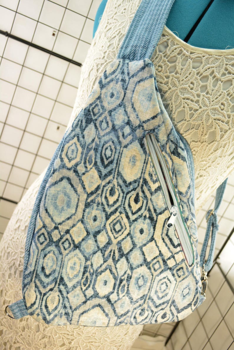 K&ouml;nnte beinhalten: Eine Sling-Tasche mit blauem und cremefarbenem geometrischem Muster mit einem Jeans-Tr&auml;ger und Rei&szlig;verschluss. Die Tasche hat ein sich wiederholendes Muster aus Sechsecken, Rauten und anderen Formen. Der Rei&szlig;verschluss ist silberfarben.
