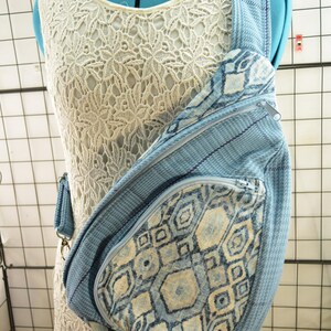 May include: A blue and white patterned sling bag with a zippered pocket and adjustable strap. The bag features a geometric design and is displayed on a mannequin wearing a white lace dress.