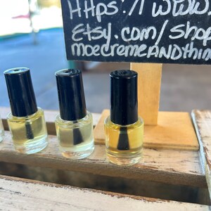 May include: Three small, clear glass bottles of liquid with black caps are displayed on a wooden surface. The bottles are filled with a yellow-tinted liquid. A chalkboard sign in the background displays the text "and Things" and a website address.