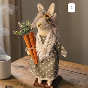 May include: A decorative Easter bunny figurine. The bunny wears a gray polka-dot dress, a white lace collar, and a burlap bow. It holds two orange carrot plushies tied with twine. The bunny is on a wooden base, perfect for Easter decor.