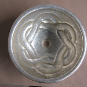 May include: A silver hammered metal sink with a decorative design of interwoven circles. The sink has a round shape and a smooth, polished finish.