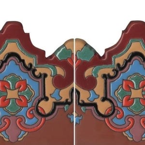 May include: Two identical decorative ceramic tiles featuring a scalloped top edge. The design displays a symmetrical pattern with deep red, light blue, tan, and dark green sections outlined in black, centered around a four-petaled floral motif.