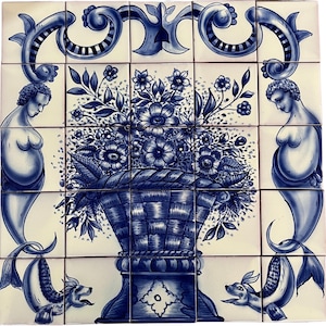 May include: A blue and white ceramic tile mural depicting a vase with a basket of flowers. The vase is decorated with a blue and white floral pattern. Two mermaids flank the vase, each with a blue tail and a white body. The tile is made up of 16 individual tiles.