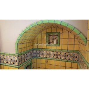 Op de afbeelding: Een gele betegelde boog met groene afwerking en een kleinere betegelde nis. De nis heeft een witte kaars erin. De tegels hebben een traditioneel Mexicaans ontwerp.