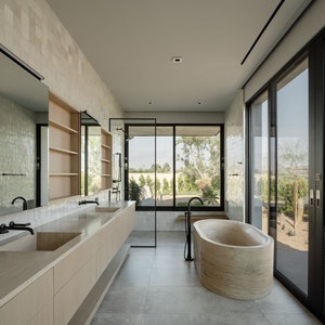 May include: A modern bathroom with a neutral color palette. Features include a large stone bathtub, double sinks, and large windows with a view of the outdoors. The walls are tiled, and the fixtures are black.