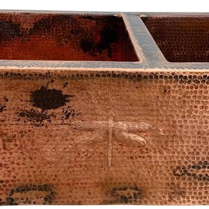 May include: A double-basin farmhouse sink crafted from hammered copper with a dark, weathered patina. The front apron features three embossed dragonfly motifs evenly spaced across the textured surface, showcasing a rich, metallic brown finish.