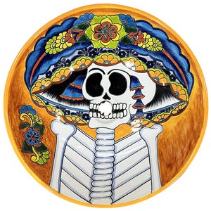 Peut inclure: Une vasque ronde en céramique peinte à la main représentant un squelette portant un grand chapeau coloré orné de motifs floraux dans des tons de bleu, jaune, orange et vert sur un fond orange chaud et texturé.