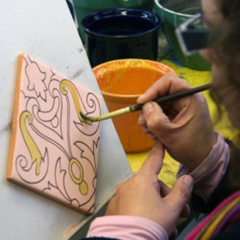 May include: A person painting a decorative ceramic tile with a brush. The tile is peach-colored with a floral design outlined in black and filled with yellow paint. Other ceramic items and paint are in the background.