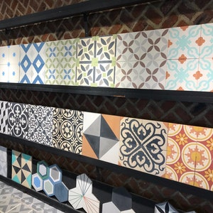 May include: A collection of decorative ceramic tiles with various geometric and floral patterns in black, white, blue, green, orange, and red colors. The tiles are arranged on a shelf with a brick wall background.