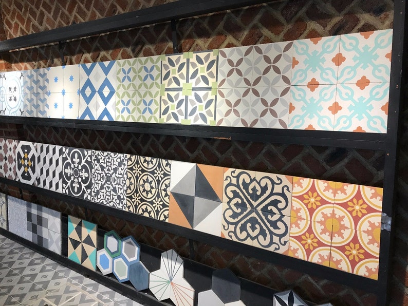 May include: A collection of decorative ceramic tiles with various geometric and floral patterns in black, white, blue, green, orange, and red colors. The tiles are arranged on a shelf with a brick wall background.