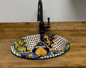Op de afbeelding: Een kleurrijk keramiek wastafel met een blauw, groen, geel en wit bloemenpatroon. De wastafel is gemonteerd op een houten aanrechtblad met een zwarte kraan.