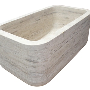 May include: A rectangular, light-colored stone bathtub with rounded corners. The tub has a smooth, polished interior and a textured exterior, showcasing the natural grain of the stone. The design is minimalist and modern, suitable for a spa-like bathroom.