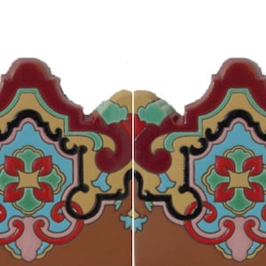 May include: A decorative ceramic tile featuring a symmetrical, ornate pattern with a scalloped top edge. The design incorporates vibrant colors including deep red, light blue, yellow, green, and black, set against a solid brown base color.