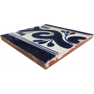 May include: Square ceramic tile with a cream-colored background and a navy blue floral design. The tile has a navy blue border with two white stripes. The edges of the tile are a reddish-brown color.