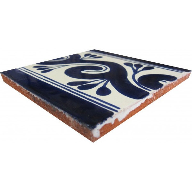 May include: Square ceramic tile with a cream-colored background and a navy blue floral design. The tile has a navy blue border with two white stripes. The edges of the tile are a reddish-brown color.