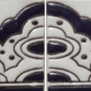May include: A rectangular ceramic tile featuring a raised, embossed black and white pattern. The design consists of a central floral motif flanked by symmetrical curved lines, dots, and scalloped edges, creating a repeating decorative border.