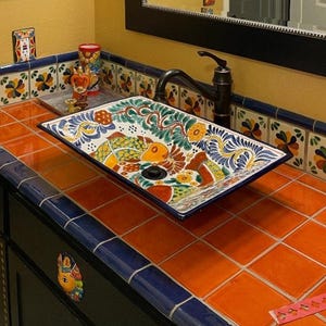May include: A colourful ceramic sink with floral and geometric designs sits on a countertop with orange and blue tiles. The sink is rectangular and has a single faucet.