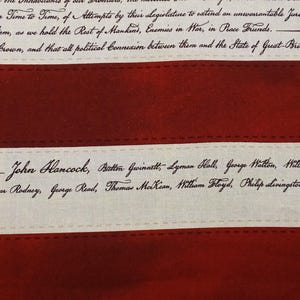 May include: Close-up of a fabric with a red and cream color scheme. The fabric features text from the Declaration of Independence, including the names of signers like John Hancock. The design resembles a flag.