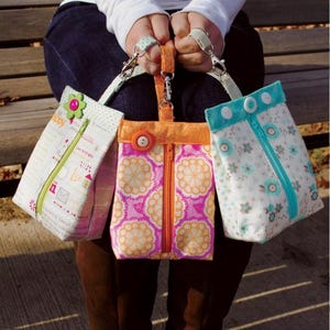 May include: Three colorful fabric tote bags with zippers and handles. The bags feature floral and patterned designs in pink, orange, and blue. Each bag has a different decorative element, including buttons and a flower.