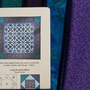 May include: A printed paper with a quilt design, featuring a geometric pattern in shades of blue, teal, and purple. The text reads "Large Economy Block" and instructions for the quilt. The background includes purple fabric with a leaf pattern and teal fabric.