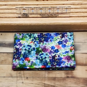 May include: A folded piece of fabric with a watercolor floral print featuring blue, purple, green, and red flowers on a white background. The fabric is labeled "Fabrics by the River".