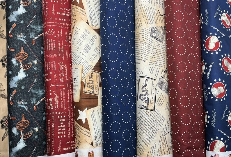May include: A collection of fabric rolls with patriotic and historical designs. Patterns include cannons, text, stars, and circles. Colors include navy, red, tan, and white. Some rolls feature text like "We the People" and "Join or Die."
