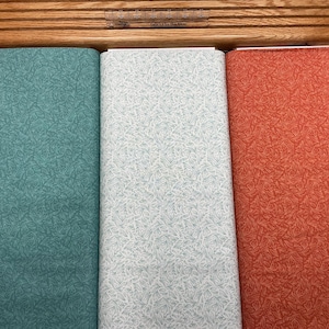 May include: Three rolls of fabric with a leaf pattern in teal, white, and orange. The fabric is displayed on a wooden cutting board with a ruler. The fabric is tightly rolled and ready for use.