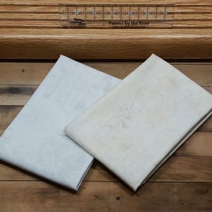 May include: Two pieces of white fabric, one with a subtle pattern and the other plain, are folded and laid on a wooden surface. The fabric is soft and smooth, and the pattern is delicate and understated.