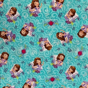 May include: A turquoise fabric with a repeating pattern of a cartoon princess in a purple dress. The princess is holding a red jewel and the text "The Power of the Amulet" is written in white. The background is a swirling turquoise pattern with white hearts and butterflies.