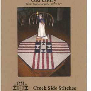 May include: A red, white, and blue table topper with a star pattern. The table topper is made of fabric and is approximately 27 inches by 27 inches. The table topper is called "Old Glory" and is pattern #157 from Creek Side Stitches.