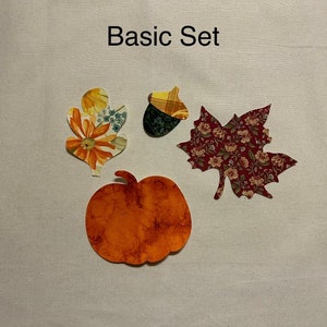 May include: Basic Set of fall-themed fabric appliques. The set includes a pumpkin, an acorn, a maple leaf, and an oak leaf. The pumpkin is orange, the acorn is brown and yellow, the maple leaf is red and white, and the oak leaf is yellow and green.