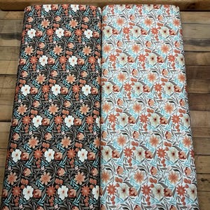 May include: Two rolls of floral fabric. One features a dark brown background with white, orange, and peach flowers. The other has a white background with orange, peach, and blue flowers. The text "Fabrics by the River" is visible.