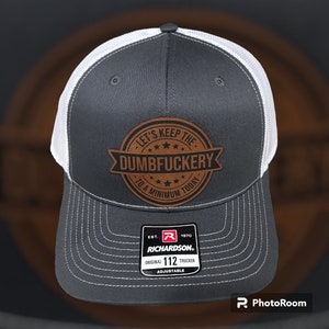 May include: A gray and white trucker hat with a brown leather patch that says "Let's Keep The Dumbfuckery To A Minimum Today". The hat is adjustable and has a Richardson 112 logo.