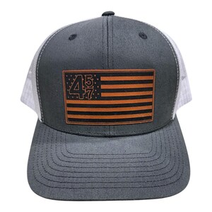 May include: A gray and white trucker hat with a brown leather patch featuring an American flag with the numbers "45 47" printed on it.
