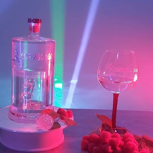 Op de afbeelding: Een heldere glazen fles Sensual Gin met een roze en groen licht dat erop schijnt. De fles staat op een wit platform met een paar rode frambozen en een heldere glazen ijsblokje. Een helder glazen gin-glas met de woorden "Sensual Gin" gegraveerd aan de zijkant staat op een tafel met een paar rode frambozen en groene bladeren.