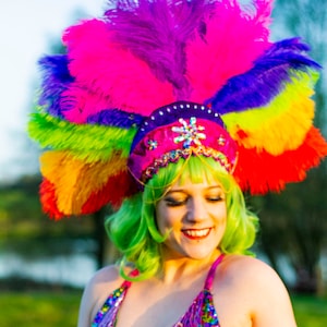May include: A vibrant, sequined halter top and matching headdress with a rainbow of feathers. The outfit features a pink, purple, and blue color scheme with fringe detailing. The model is wearing a green wig.