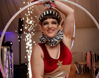 May include: A woman wearing a red and white striped costume with a gold star and a black and white striped collar is performing with a hoop. She is smiling and has black and white makeup on her eyes.