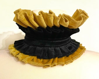 Può includere: Un colletto a balze a strati in velluto oro e nero. Il colletto è esposto su un manichino, mettendo in mostra i colori e le trame contrastanti. Il design presenta più strati di tessuto, creando un look drammatico ed elegante.