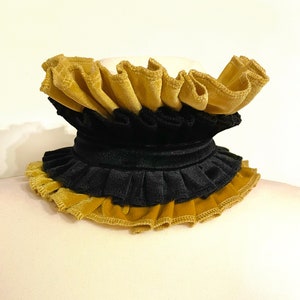 May include: A layered, ruffled collar in gold and black velvet. The collar is displayed on a mannequin, showcasing the contrasting colors and textures. The design features multiple tiers of fabric, creating a dramatic and elegant look.
