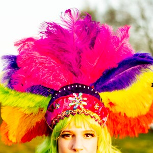 May include: A vibrant, multi-colored feather headdress. The headdress features a rainbow of colors, including pink, purple, yellow, green, and orange. The base is red with decorative embellishments. The image is brightly lit, with a soft focus background.