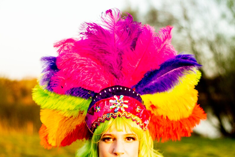 May include: A vibrant, multi-colored feather headdress. The headdress features a rainbow of colors, including pink, purple, yellow, green, and orange. The base is red with decorative embellishments. The image is brightly lit, with a soft focus background.