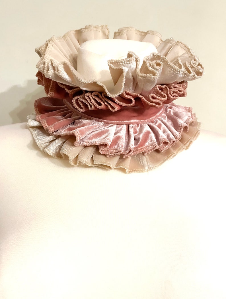 Peut inclure: Un col &agrave; volants superpos&eacute;s dans les tons cr&egrave;me, rose blush et rose. Le col pr&eacute;sente plusieurs niveaux de tissu pliss&eacute; et une bande de velours, cr&eacute;ant un design textur&eacute; et orn&eacute;. Le col est probablement un accessoire de costume.