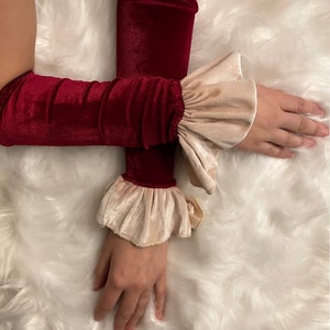 May include: Burgundy velvet arm warmers with cream-colored ruffled cuffs. The arm warmers are made of a soft, plush material and are designed to cover the arms from the wrist to the elbow. A unique fashion accessory.