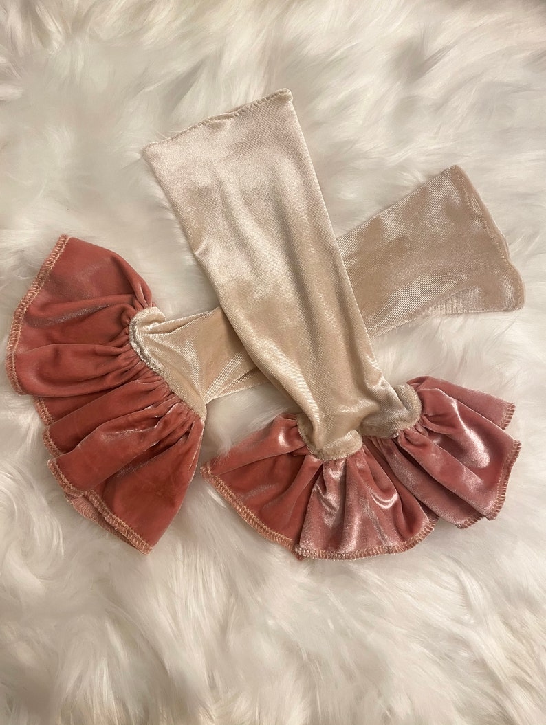 May include: A pair of velvet arm warmers with a ruffled trim. One arm warmer is beige and the other is a dusty rose color.