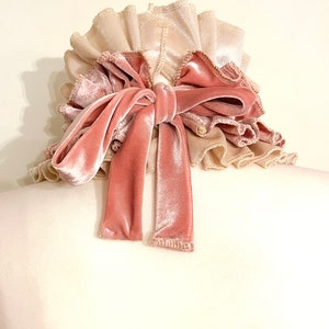 Peut inclure: Un chouchou &agrave; plusieurs couches avec une texture douce et velout&eacute;e. Le chouchou pr&eacute;sente une base beige clair avec des bords fronc&eacute;s, recouverte d'un n&oelig;ud en velours rose poudr&eacute;. Les longues queues fluides du n&oelig;ud ajoutent une touche d'&eacute;l&eacute;gance.