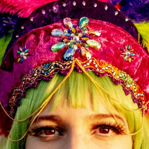 May include: A close-up of a colorful headdress with a magenta velvet crown adorned with gemstones and gold trim. The headdress is decorated with a rainbow of feathers. The person has green hair and red eyes.