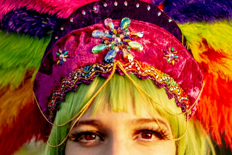 May include: A close-up of a colorful headdress with a magenta velvet crown adorned with gemstones and gold trim. The headdress is decorated with a rainbow of feathers. The person has green hair and red eyes.