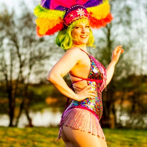 May include: A vibrant burlesque costume featuring a sequined bodysuit with fringe, a colorful feathered headdress, and green wig. The costume is predominantly pink, with accents of purple, yellow, and blue. The outfit is designed for stage performance.